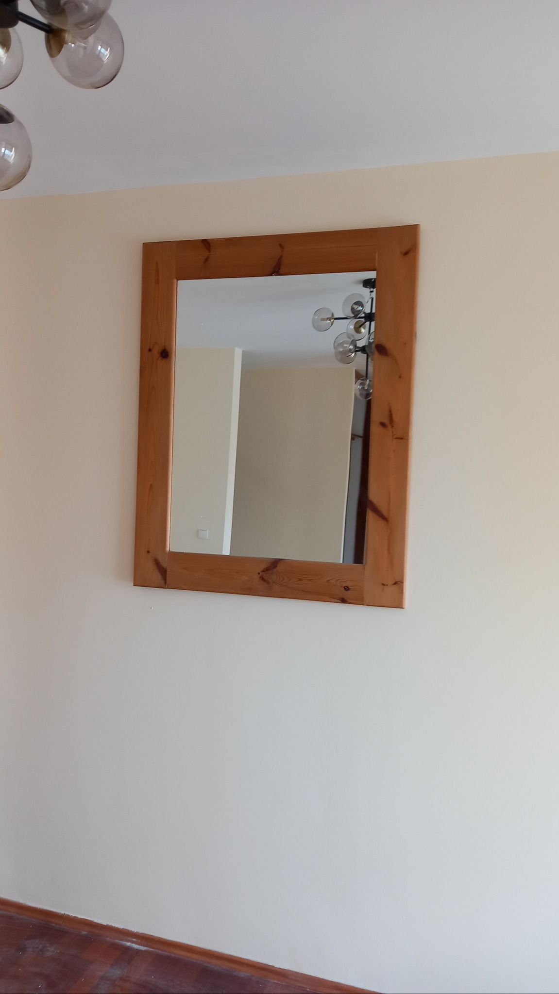 Kwadratowe lustro w drewnianej ramie wisi na kremowej ścianie. W odbiciu widać fragment sufitu z nowoczesną lampą i białą ścianę.