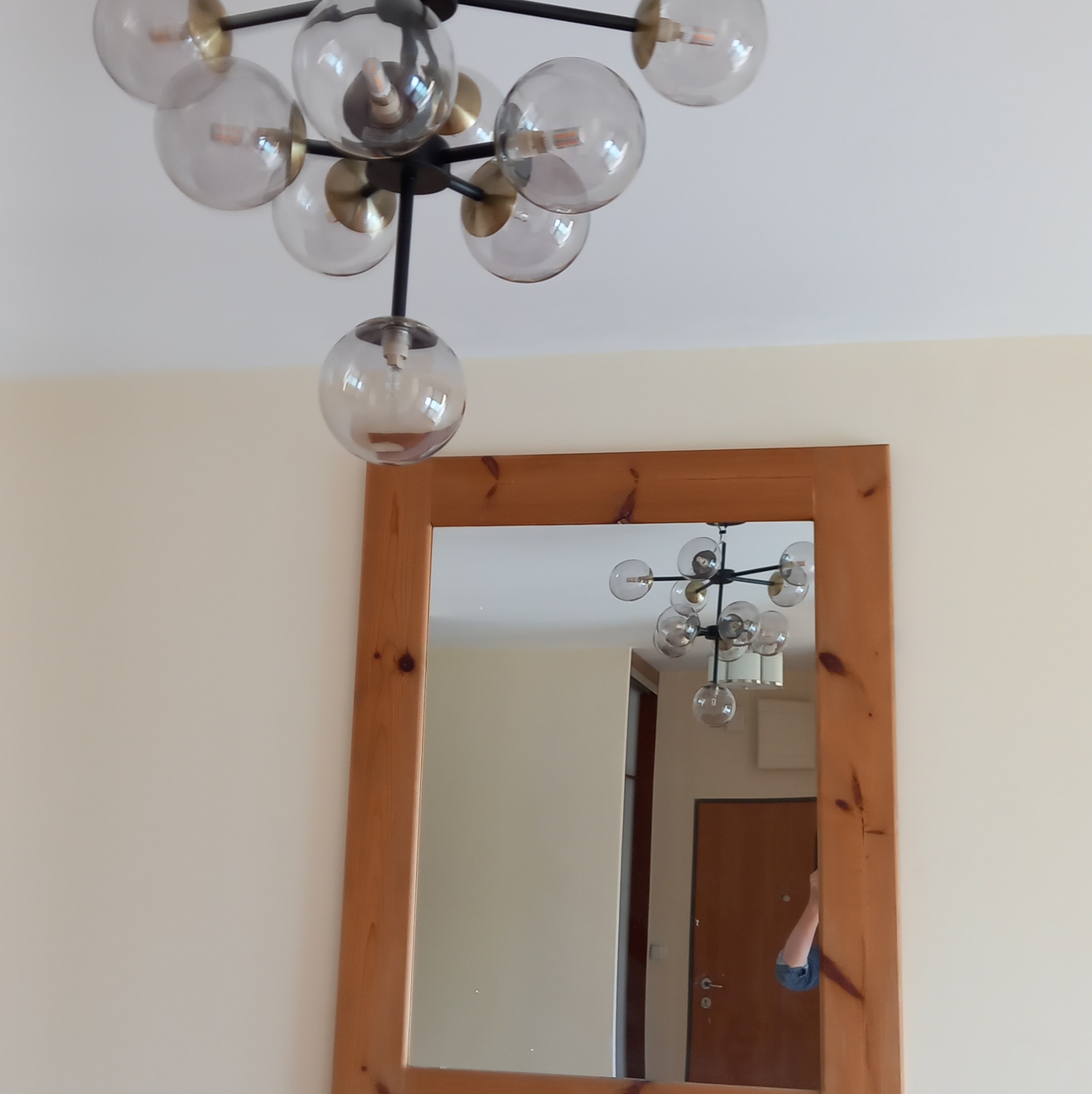 Nowoczesna lampa sufitowa z kulistymi kloszami, odbijająca się w lustrze z drewnianą ramą na kremowej ścianie. Widok z dołu, oświetlenie wnętrza.