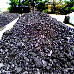 Góra błyszczącego ekogroszku na składzie, z workami w tle i zielonymi drzewami. Ujęcie z dołu podkreśla fakturę i ilość węgla.