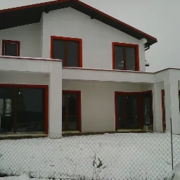 Nowoczesny dom jednorodzinny z białą elewacją i czerwonymi obramowaniami okien, częściowo ośnieżony, widok zza metalowej siatki ogrodzeniowej.