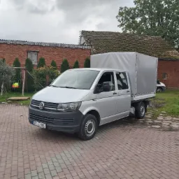Srebrny Volkswagen Transporter z plandeką na tle ceglanego budynku gospodarczego z uszkodzonym dachem, stojący na brukowanej nawierzchni.