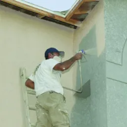 Pracownik w masce ochronnej, stojący na drabinie, aplikuje natryskowo farbę na elewację budynku z teksturą, używając agregatu.