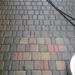 Kostka brukowa po myciu ciśnieniowym, widoczna różnica czystości, fragment ściany z cegły i dysza myjąca.