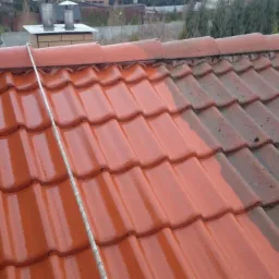 Częściowo umyty dach z czerwonej dachówki. Połowa dachu jest czysta i mokra, druga połowa brudna i sucha. Widoczna lanca do mycia.