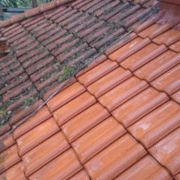 Częściowo oczyszczony dach z czerwonej dachówki ceramicznej, lewa strona pokryta mchem i zabrudzeniami, prawa strona świeżo umyta, widoczny przewód doprowadzający wodę.