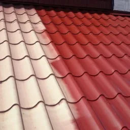 Częściowo pomalowany dach z blachodachówki w kolorze czerwonym, widoczny etap malowania z odróżniającą się niepomalowaną częścią w kolorze kremowym.