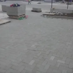 Ułożona szara kostka brukowa w jodełkę na placu budowy. Na paletach ułożone kostki. Widoczny fragment chodnika z obrzeżami i ziemią. W tle budynek.