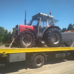Czerwony traktor z kabiną zabezpieczony pasami na żółtej platformie pojazdu pomocy drogowej z białym nadwoziem na tle błękitnego nieba i zieleni.