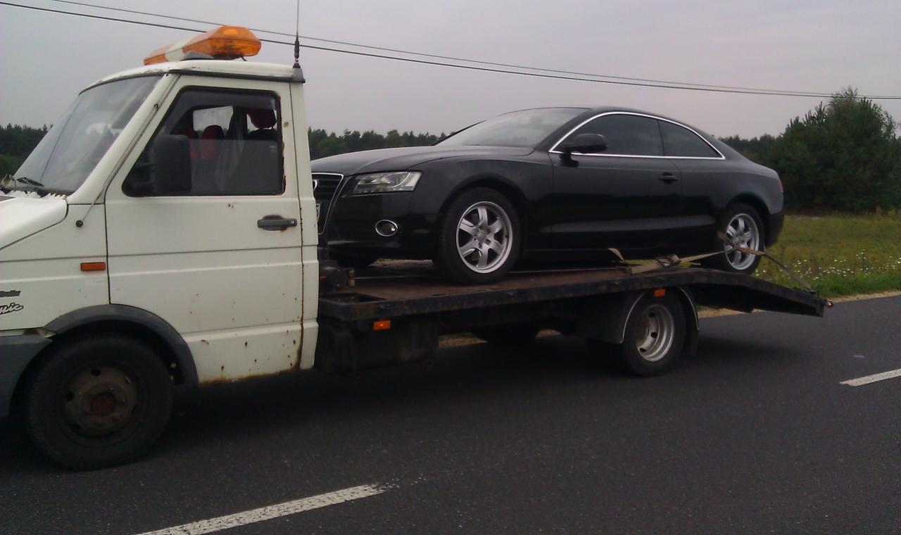 Biała laweta z pomarańczowym światłem przewozi czarne Audi A5 na platformie. Widok z boku na tle drogi i zieleni.