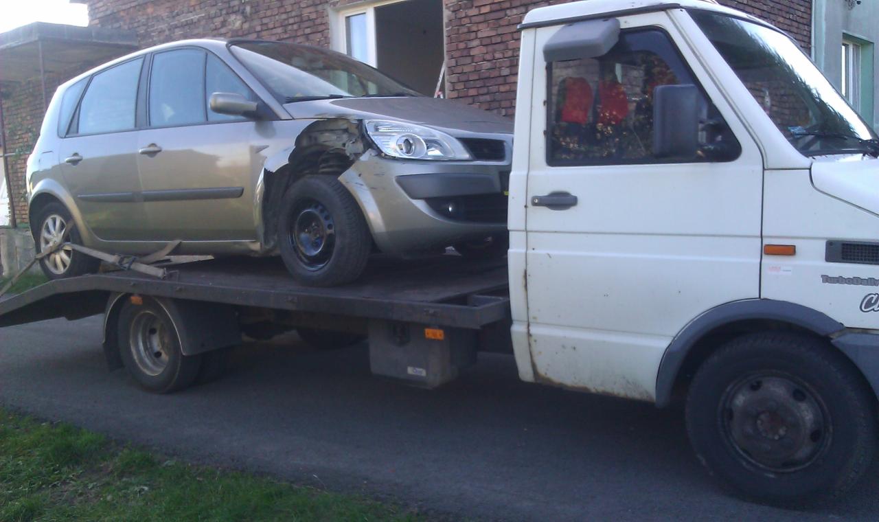 Uszkodzony, srebrny samochód osobowy na platformie lawety, zabezpieczony pasami transportowymi. Widoczny budynek w tle i fragment białego pojazdu lawety.