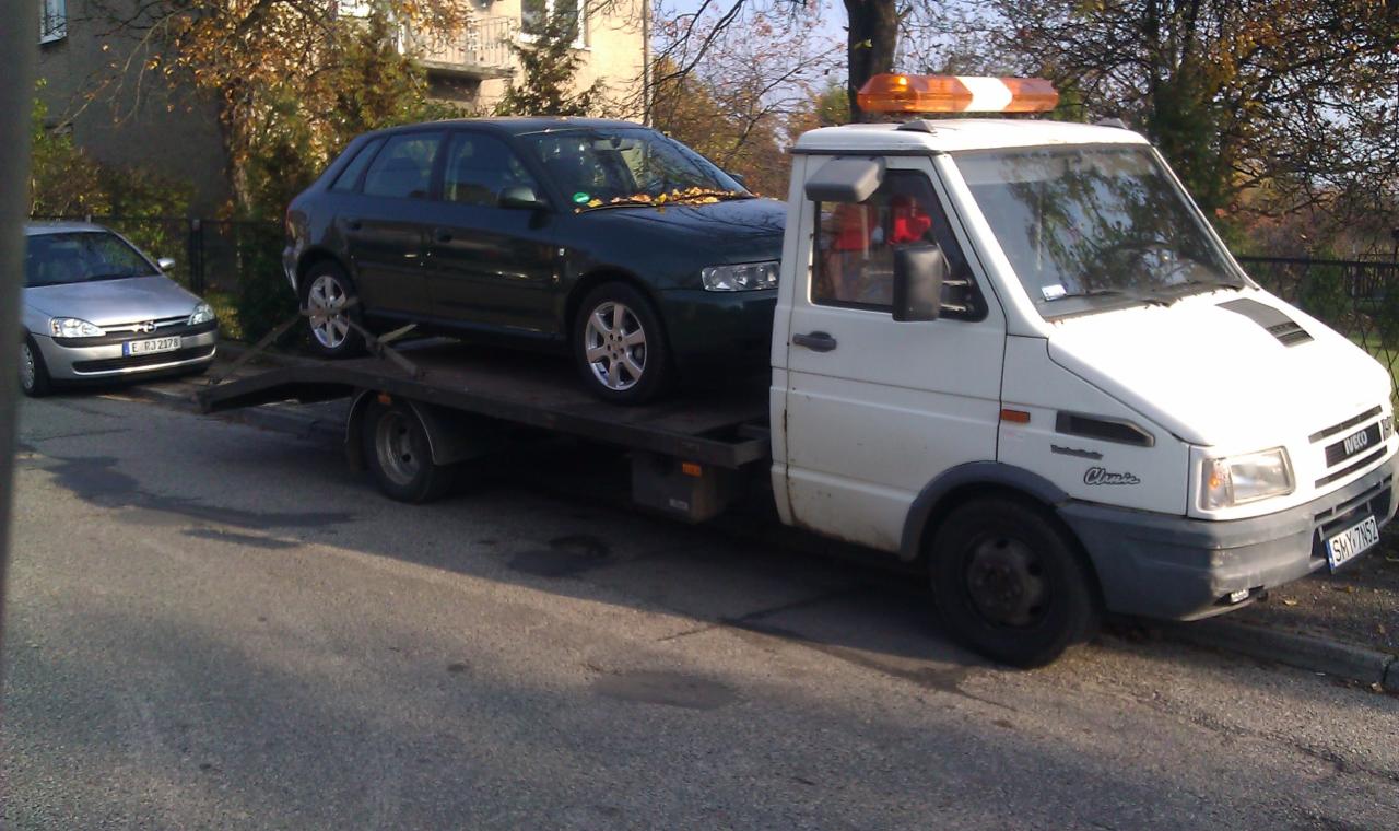 Biała laweta Iveco z pomarańczowym kogutem transportuje ciemnozielony samochód osobowy marki Audi na tle jesiennego krajobrazu. Widoczny fragment drugiego pojazdu.