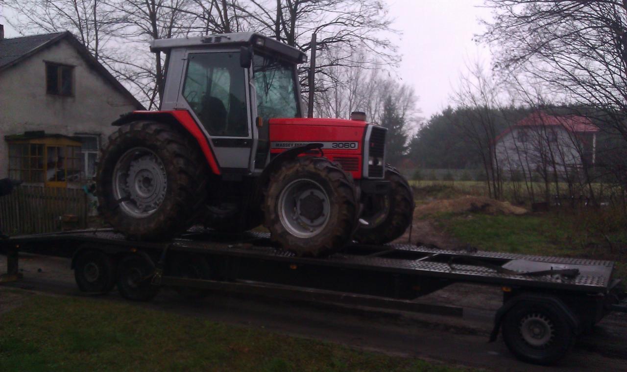 Czerwony traktor Massey Ferguson 3060 na czarnej lawecie w wiejskim otoczeniu. Widok z boku, pochmurny dzień.