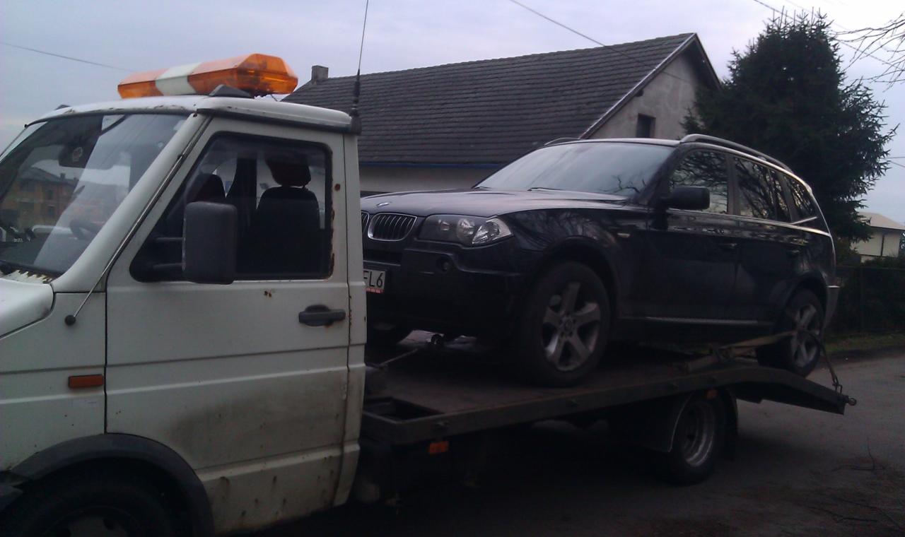 Czarny SUV BMW na platformie lawety z pomarańczowym kogutem na dachu, na tle budynków i drzew. Widok z boku, pochmurne niebo.