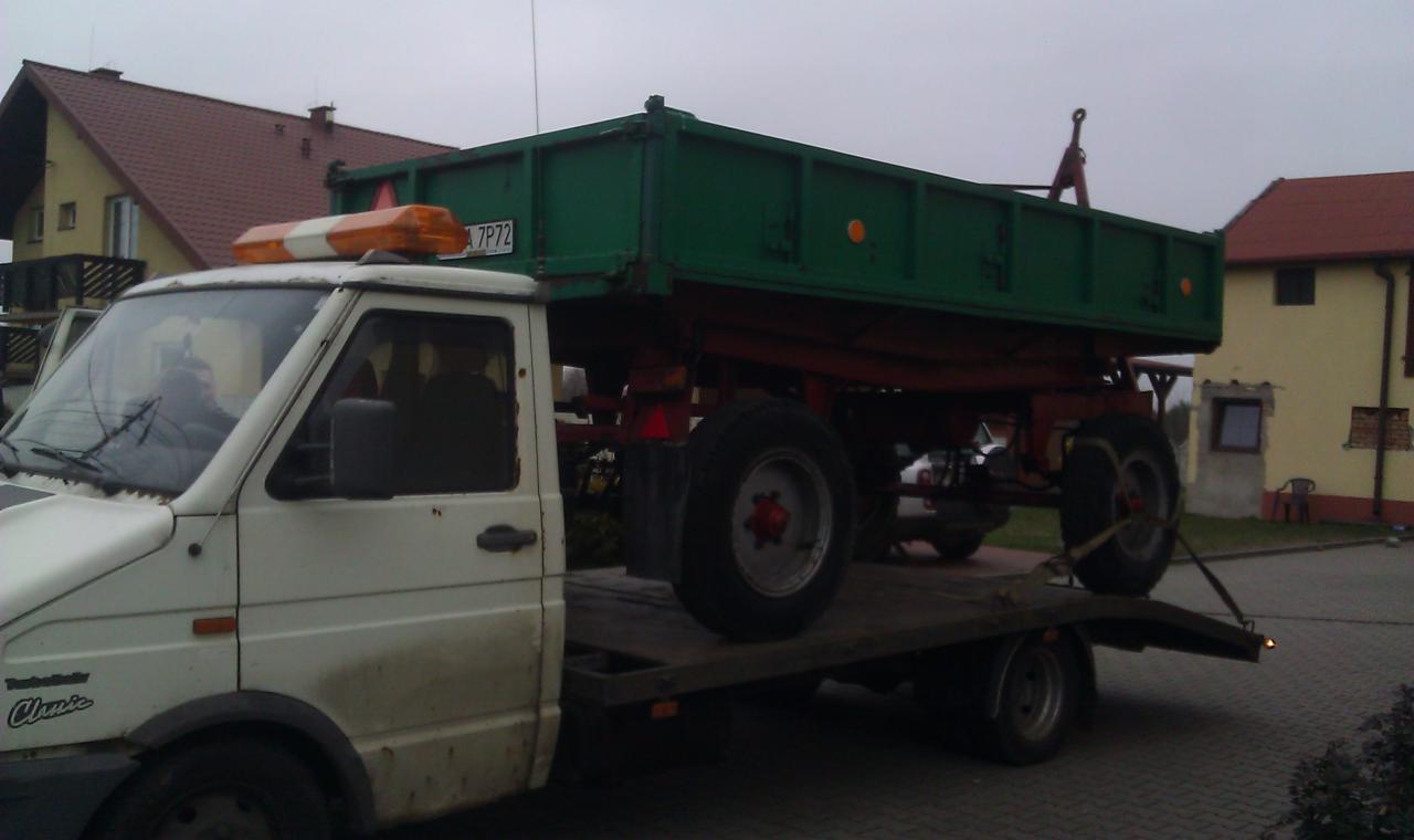 Biała laweta przewozi zieloną przyczepę rolniczą z numerem 7P72. Pomarańczowy kogut na dachu lawety. W tle budynki i pochmurne niebo.