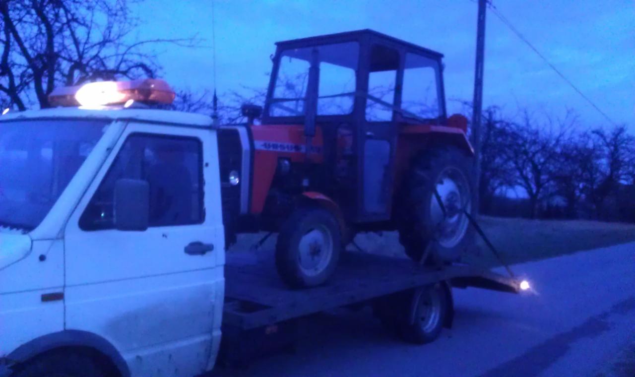 Laweta przewozi czerwony ciągnik Ursus wczesnym rankiem. Pomarańczowe światło ostrzegawcze na dachu lawety. Widoczne drzewa w tle.