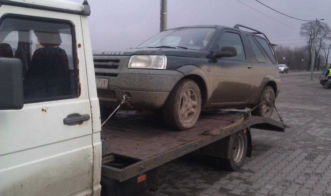 Brudny Land Rover Freelander transportowany na starej, metalowej lawecie. Widoczny fragment białej kabiny lawety i fragment chodnika z kostki brukowej.