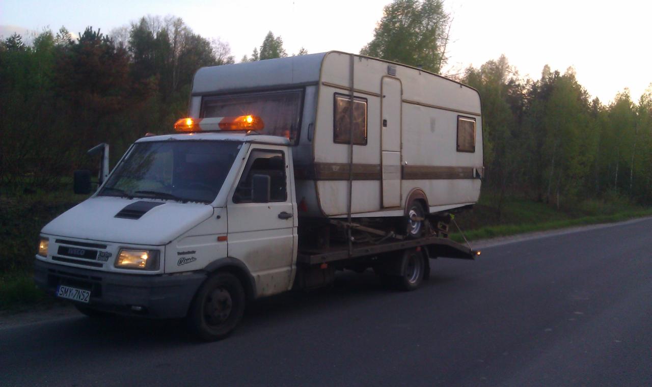 Laweta Iveco z włączonymi światłami przewozi przyczepę kempingową na platformie. Widoczne drzewa w tle i asfaltowa droga.