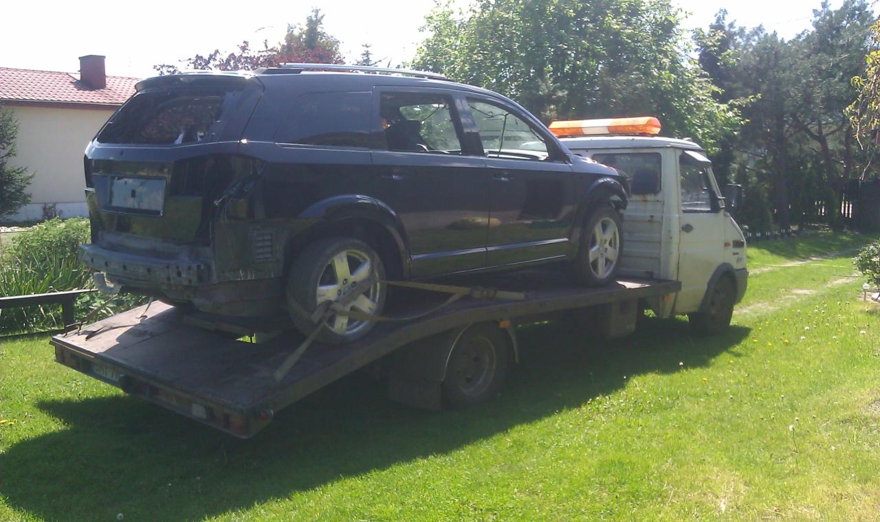 Uszkodzony, czarny SUV przewożony na platformie lawety na tle zielonego trawnika i drzew. Widoczny brak tylnej klapy i uszkodzenia karoserii.