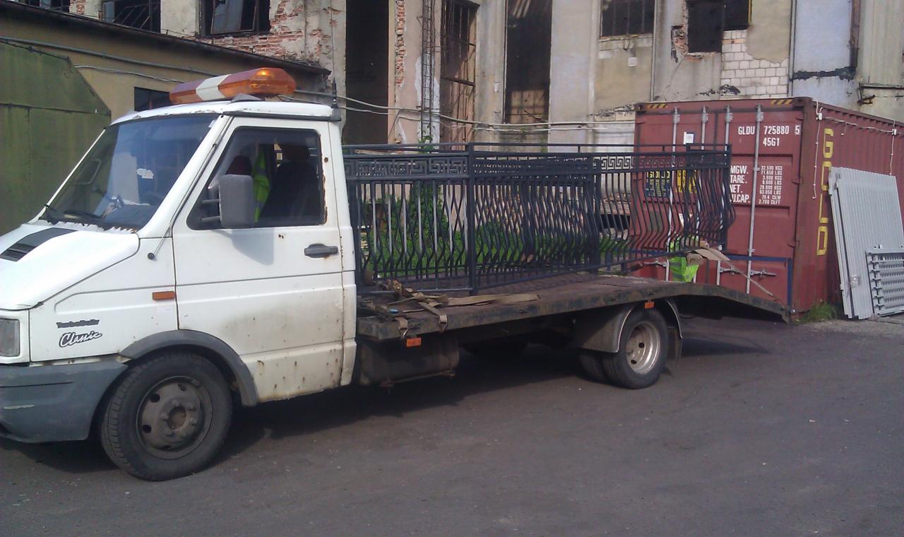 Biała laweta z pomarańczowym światłem przewozi czarne, metalowe ogrodzenie na tle budynku i kontenera. Widoczne pasy zabezpieczające ładunek.