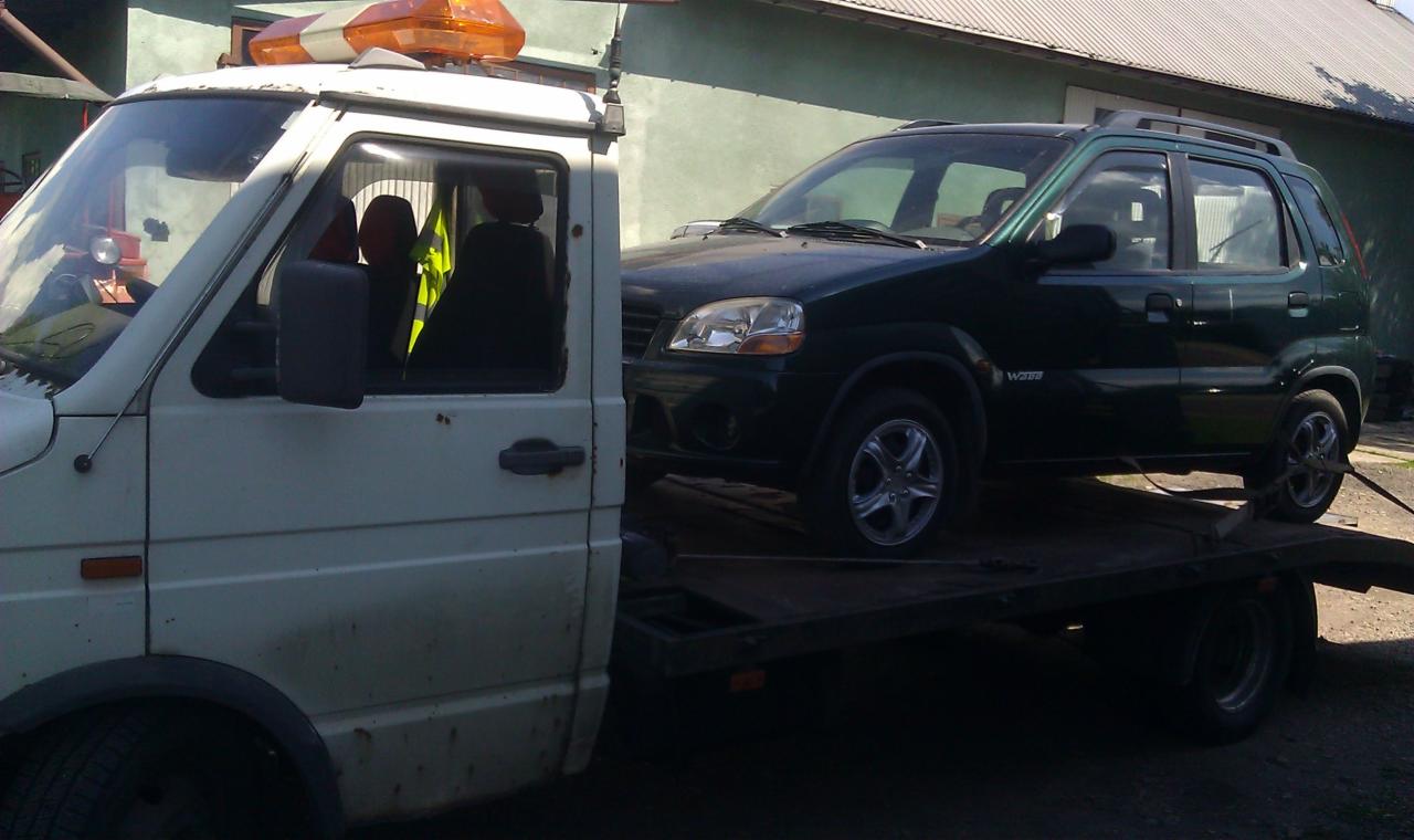 Laweta z pomarańczowym kogutem przewozi zielony samochód osobowy. Widoczny bok pojazdu na platformie. Budynek w tle.