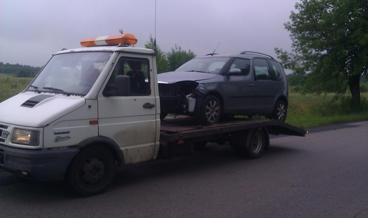 Biała laweta z uszkodzonym, szarym autem osobowym na platformie. Widoczne zielone drzewa w tle, pochmurne niebo.