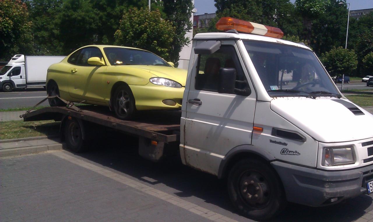 Laweta z żółtym Hyundaiem Coupe na platformie, zaparkowana na ulicy. Widoczny biały samochód dostawczy w tle i drzewa.