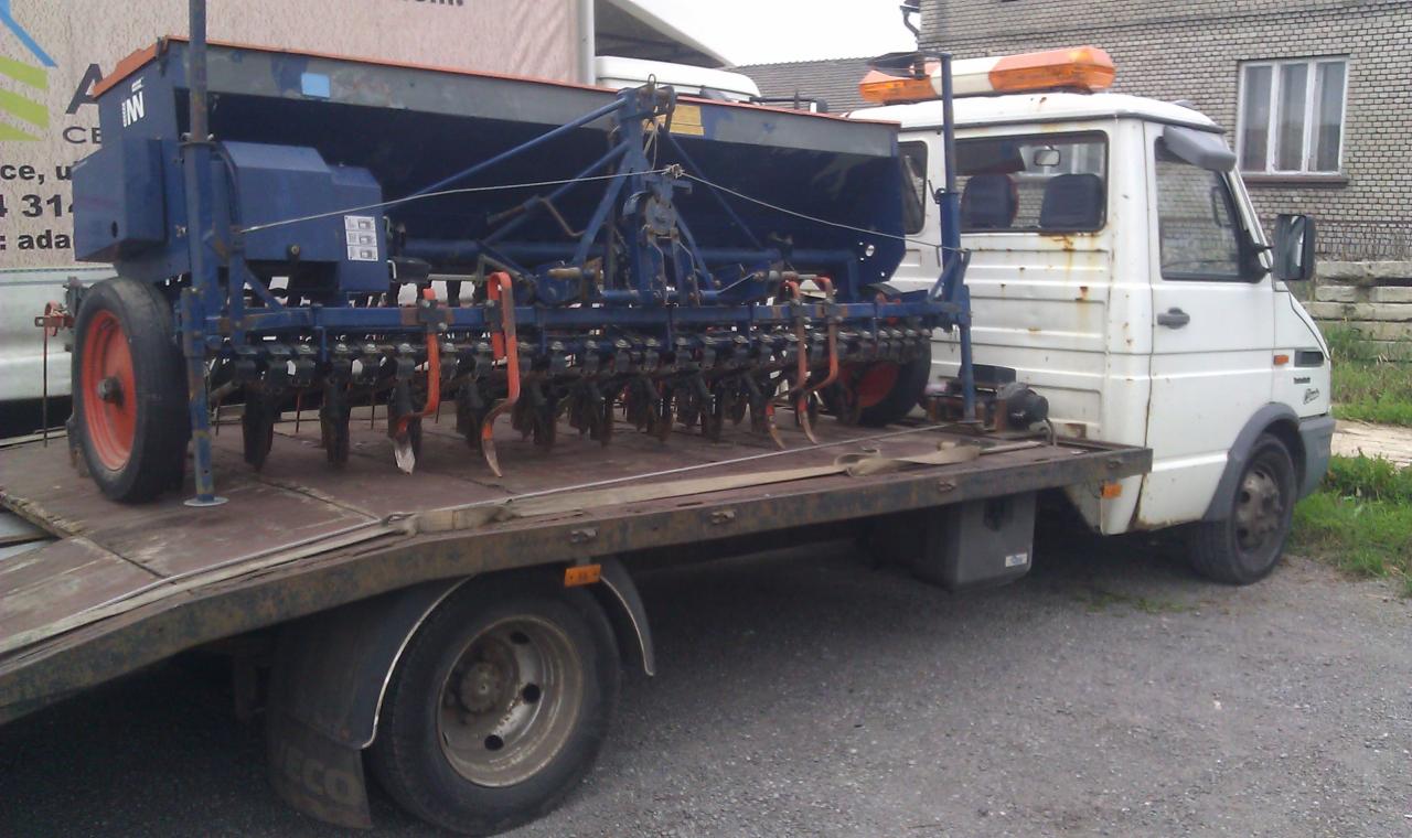 Niebieski siewnik rolniczy z pomarańczowymi elementami transportowany na platformie białego, lekko zardzewiałego samochodu dostawczego z pomarańczowym kogutem na dachu.