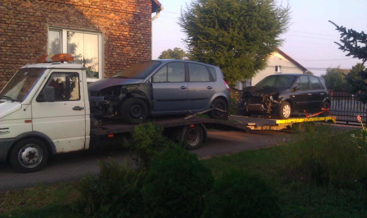 Biała laweta z pomarańczowym kogutem przewozi dwa uszkodzone samochody osobowe na tle ceglanego domu i zieleni.