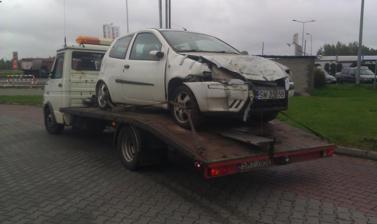 Biały uszkodzony samochód osobowy marki Fiat Punto z widoczną tablicą rejestracyjną SW 25658, zabezpieczony pasami, przewożony na platformie niskopodwoziowej w pochmurny dzień.