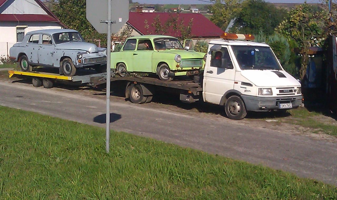 Biała ciężarówka z pomarańczowymi światłami ostrzegawczymi transportuje na lawecie dwa stare samochody: szary model GAZ-21 Wołga i zielony Trabant 601.