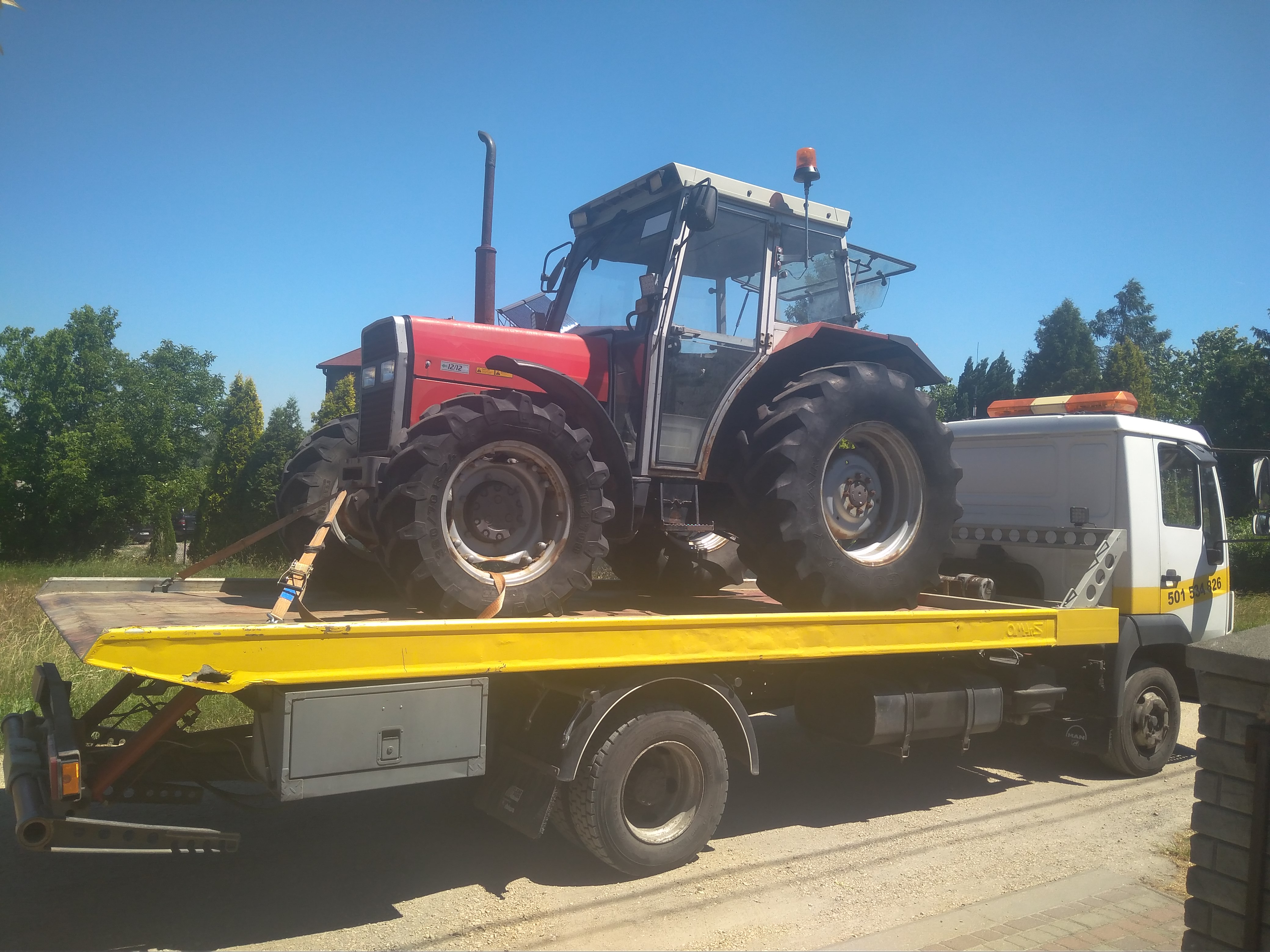 Czerwony traktor z kabiną zabezpieczony pasami na żółtej platformie pojazdu pomocy drogowej z białym nadwoziem na tle błękitnego nieba i zieleni.