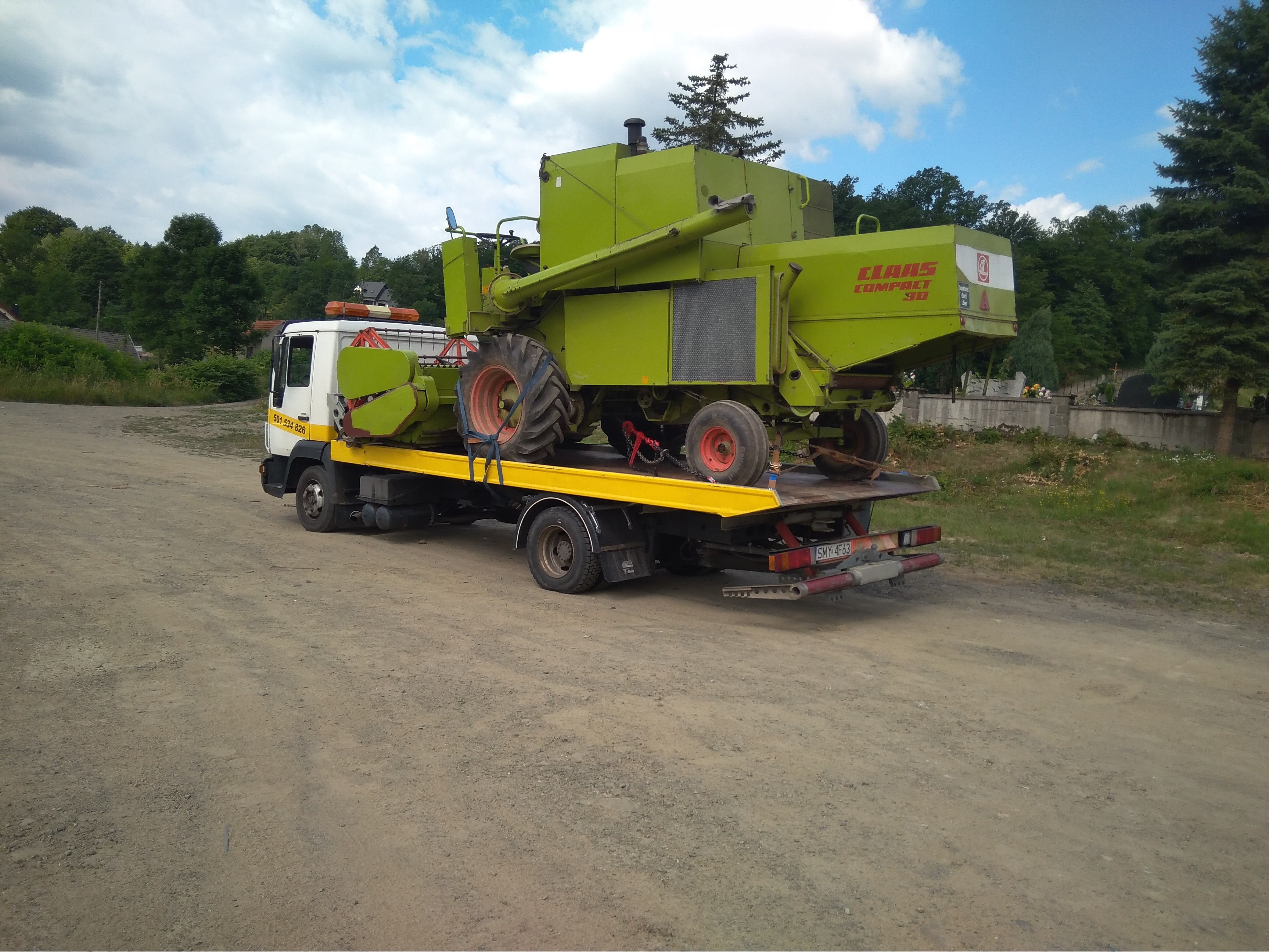 Laweta przewozi zielony kombajn Claas Compact 30 na żółtej platformie, widoczne tablice rejestracyjne i numery kontaktowe na kabinie.