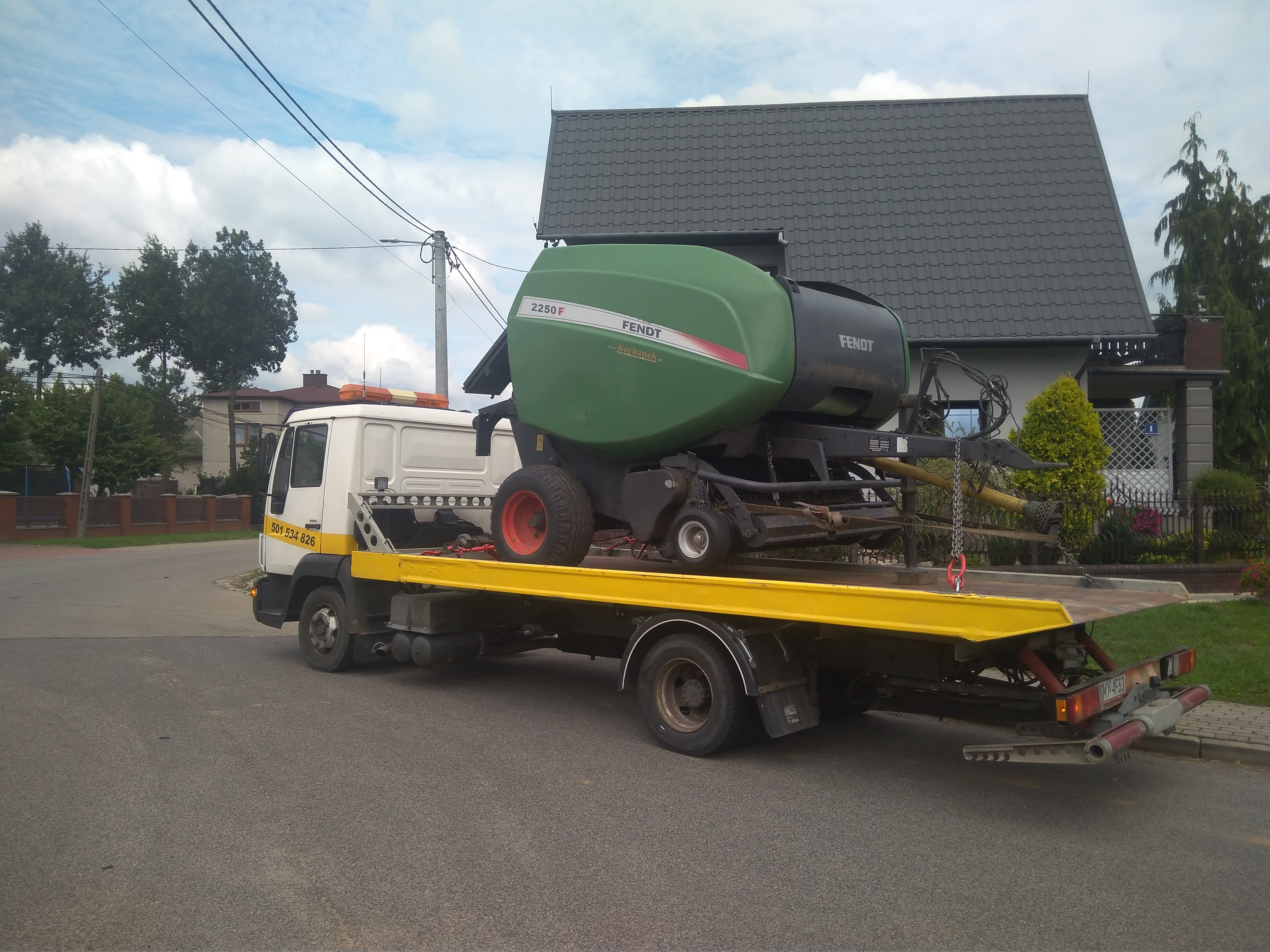 Laweta z żółtą platformą transportuje zielono-czarną prasę rolniczą Fendt 2250 F na tle domu jednorodzinnego i drzew