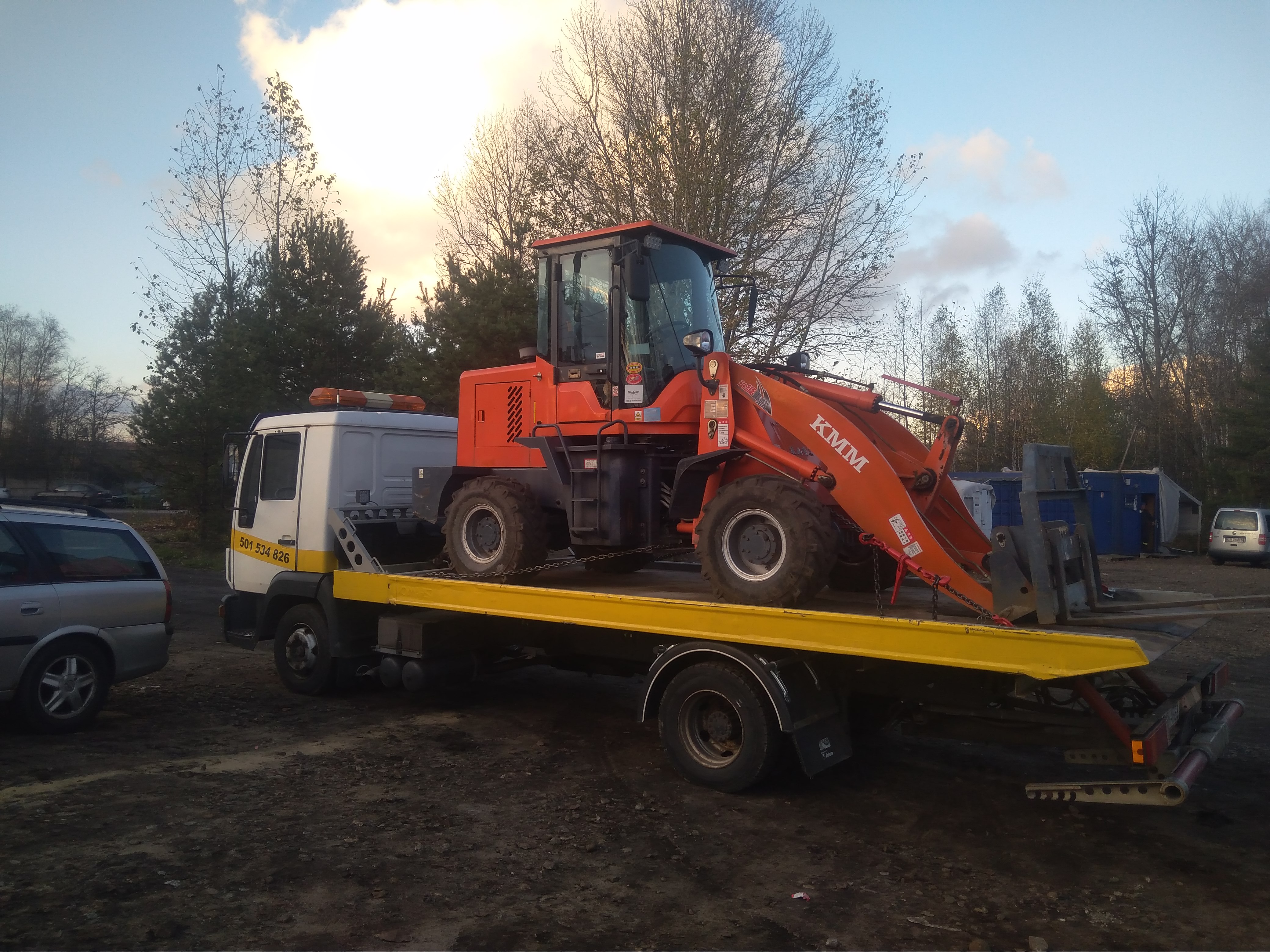 Pomarańczowa ładowarka KMM zabezpieczona łańcuchami na żółtej platformie lawety, widok z boku na tle drzew i pochmurnego nieba.