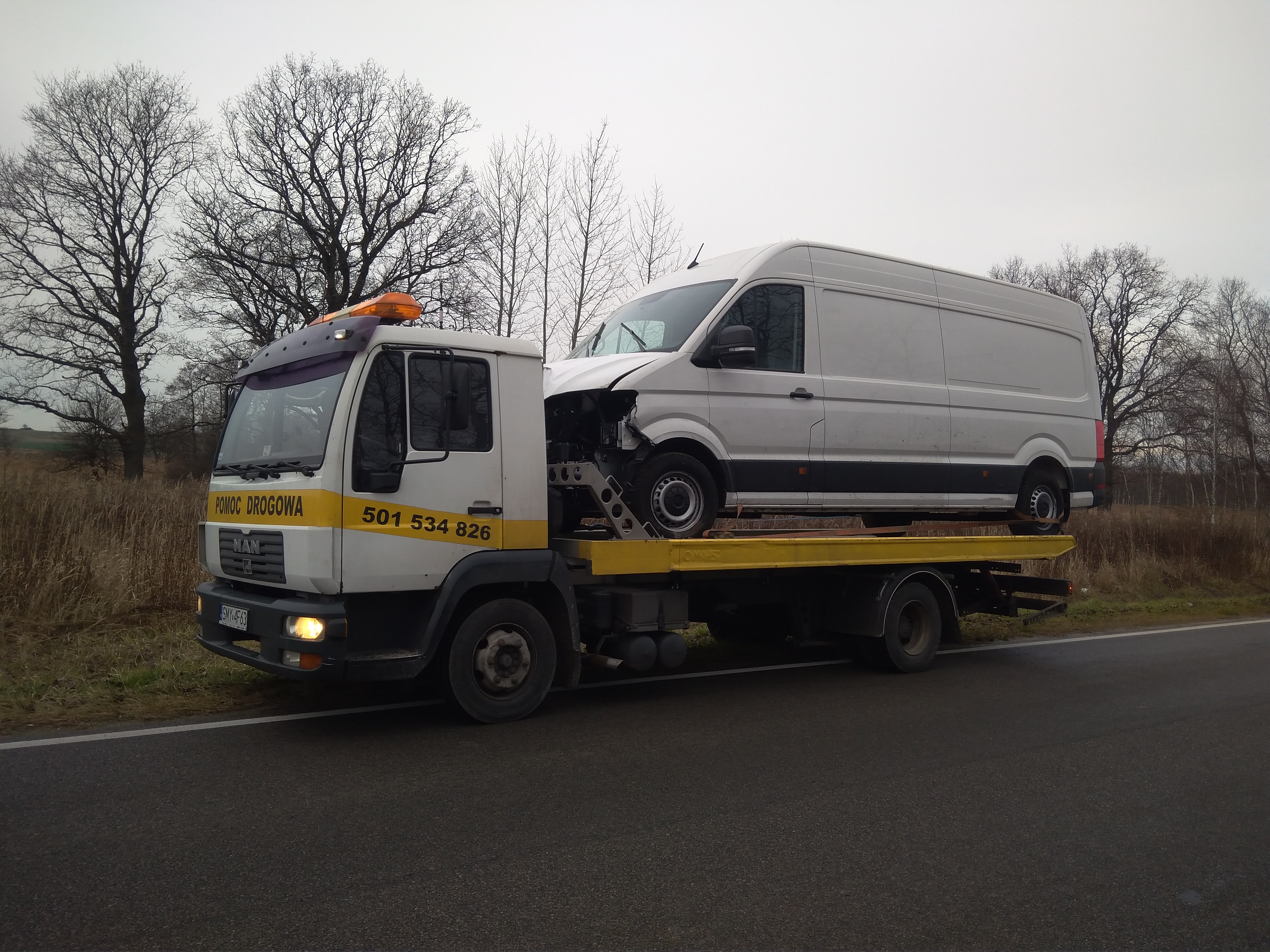 Biała laweta MAN z żółtym pasem i numerem telefonu przewozi uszkodzonego białego busa na tle szarego nieba i drzew bez liści.