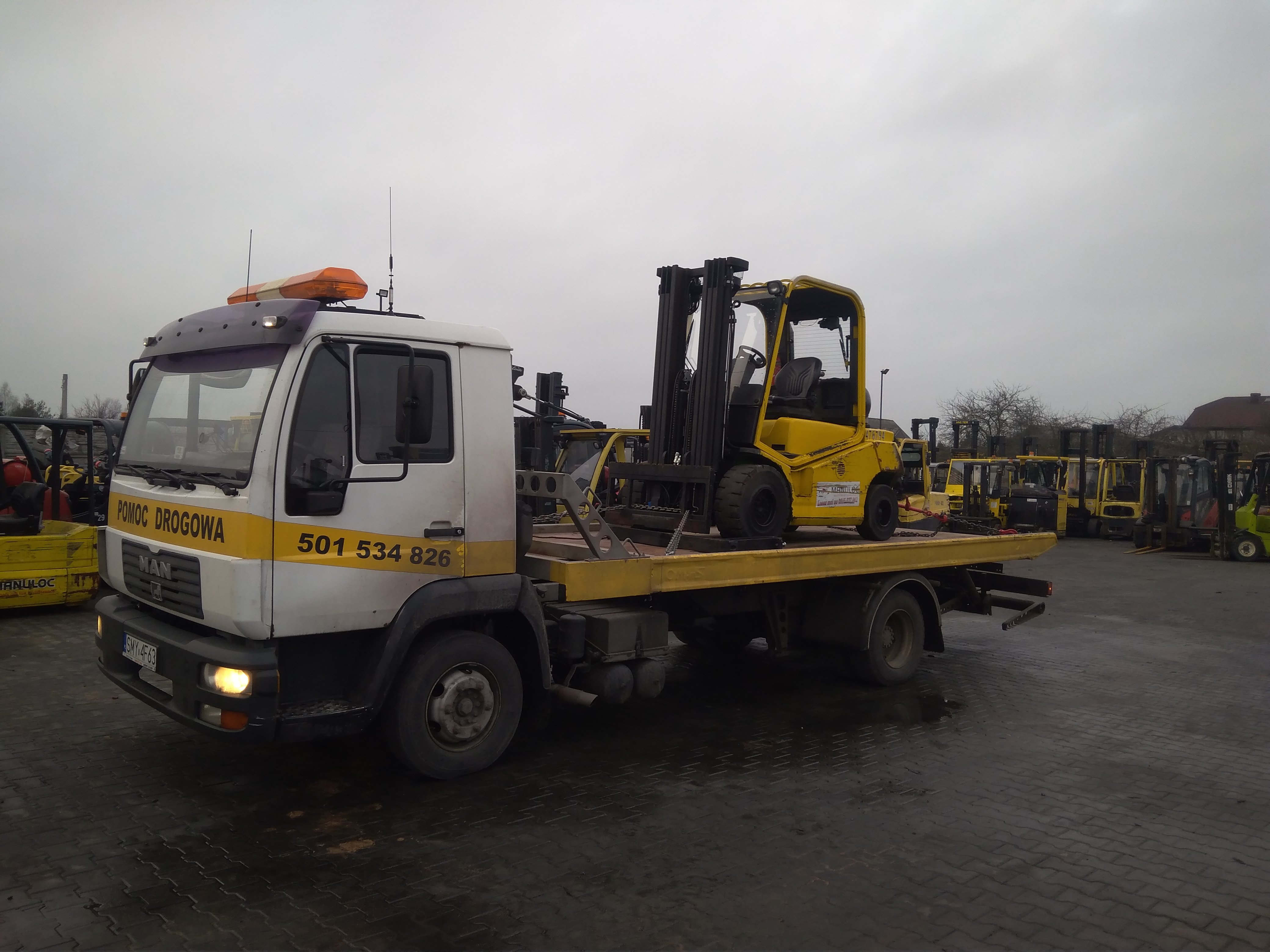 Biała ciężarówka z napisem 'Pomoc Drogowa' i numerem telefonu przewozi żółty wózek widłowy na platformie. W tle zaparkowane liczne wózki widłowe.