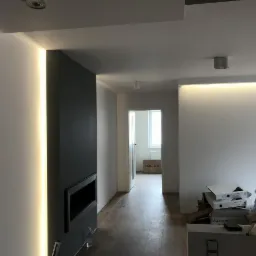 Nowoczesne wnętrze z oświetleniem LED w pionowej wnęce ściany oraz podwieszanym suficie, widoczne kartony i butelka wody na podłodze.