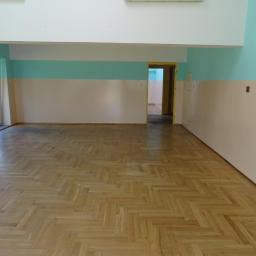 Professional Job Usługi Remontowe-Budowlane - Puste pomieszczenie z parkietem ułożonym w jodełkę, ścianami pomalowanymi w poziome pasy kremu i turkusu, z żółtą futryną drzwi prowadzących do kolejnego pomieszczenia.