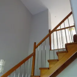 Klatka schodowa z drewnianymi schodami i metalowymi barierkami, widok z dołu, ściany pomalowane na szaro, w tle okno z luksfer&oacute;w.