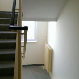 Professional Job Usługi Remontowe-Budowlane - Klatka schodowa z szarymi schodami i jasnymi ścianami. Widoczne okno z białymi ramami i grzejnik. Ujęcie z dołu, na pierwszym planie fragment poręczy.
