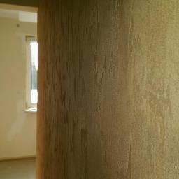 Professional Job Usługi Remontowe-Budowlane - Beżowa ściana z dekoracyjną fakturą w wykończonym pokoju, widok z bliska. W tle okno i fragment ściany przygotowanej do malowania.