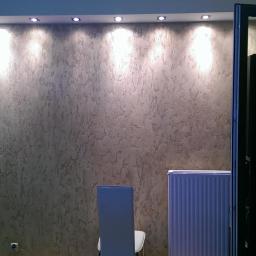 Professional Job Usługi Remontowe-Budowlane - Ściana z dekoracyjną strukturą i oświetleniem punktowym, widoczny grzejnik i krzesło. Wykończenie wnętrza w odcieniach szarości z otwartymi drzwiami w tle.