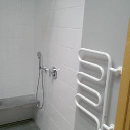 Professional Job Usługi Remontowe-Budowlane - Nowoczesna łazienka z białymi płytkami, szarymi ścianami i podłogą. Widoczny grzejnik drabinkowy, deszczownica i minimalistyczna ławka.