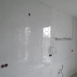 Professional Job Usługi Remontowe-Budowlane - Ściana wyłożona błyszczącymi, heksagonalnymi, białymi płytkami. Widoczne otwory na gniazdka. Fragment ściany z niedokończonymi pracami wykończeniowymi.