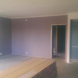 Professional Job Usługi Remontowe-Budowlane - Pokój w trakcie prac wykończeniowych: ściany pomalowane na fioletowo, podłoga surowa, widoczne płyty podłogowe w paczkach, zabudowa kuchenna w tle.