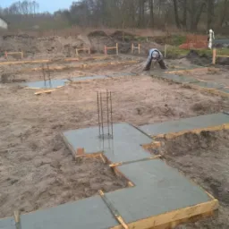 Wylane fundamenty pod budynek, widoczne szalunki i wystające zbrojenie, pracownik w oddali wyrównuje powierzchnię betonu.