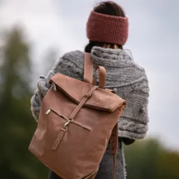 Kobieta z tyłu, ubrana w szary sweter i brązową opaskę, nosząca brązowy skórzany plecak z metalowymi detalami na tle rozmytej zieleni.