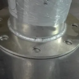 Precyzyjny spaw metalu: okrągła flansza z otworami, przyspawana do rury, widoczny ślad spawu na błyszczącej powierzchni metalu, rura owinięta folią.