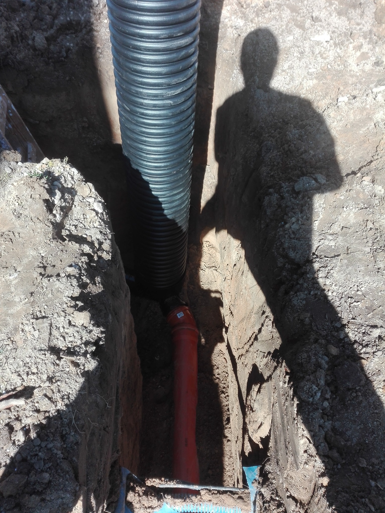 Wykop z ułożonymi rurami kanalizacyjnymi: karbowana czarna i pomarańczowa, cień sylwetki osoby na ścianie wykopu.