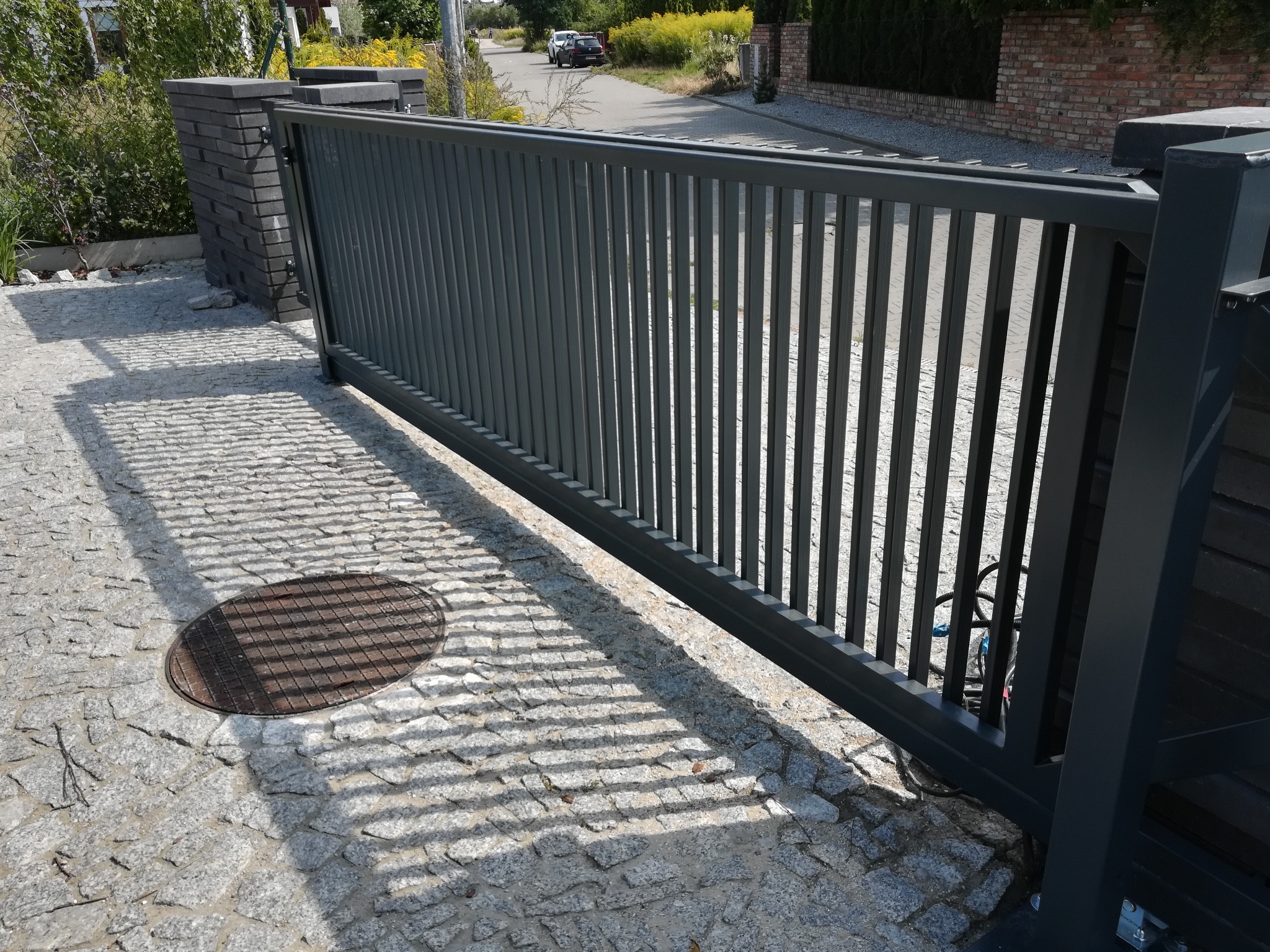 Automatyczna brama przesuwna z szarymi słupkami z cegły, na podjeździe z kostki brukowej, widoczne kable i rury od automatyki bramy.